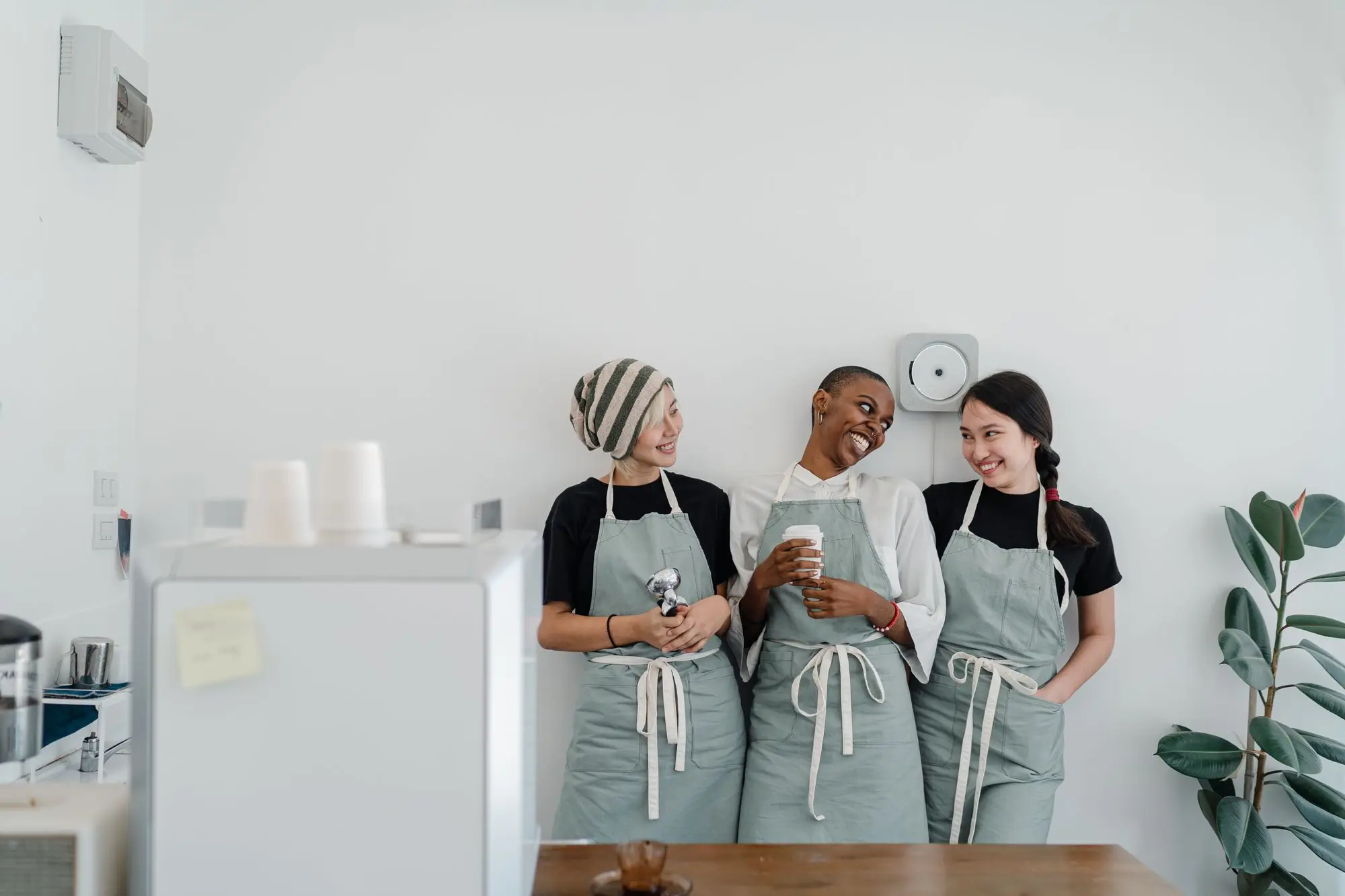Three cafe staff in matching sage aprons sharing a laugh behind the counter of a bright, minimal coffee shop